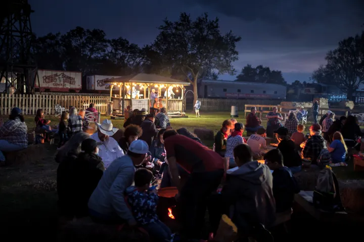 People gather around campfires at an outdoor evening event, with a lit pavilion in the background.