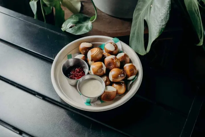 Bowl of pretzel bites with two sauces on a black table, surrounded by green leaves.