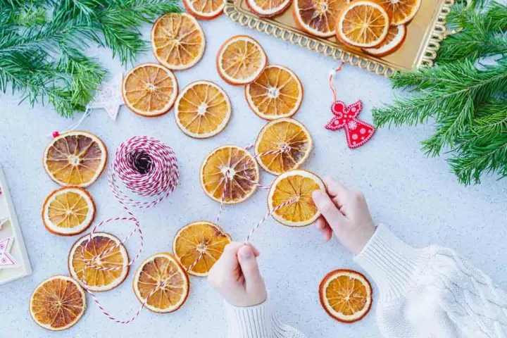 Hands stringing dried orange slices with red and white twine, surrounded by greenery and a red angel ornament.
